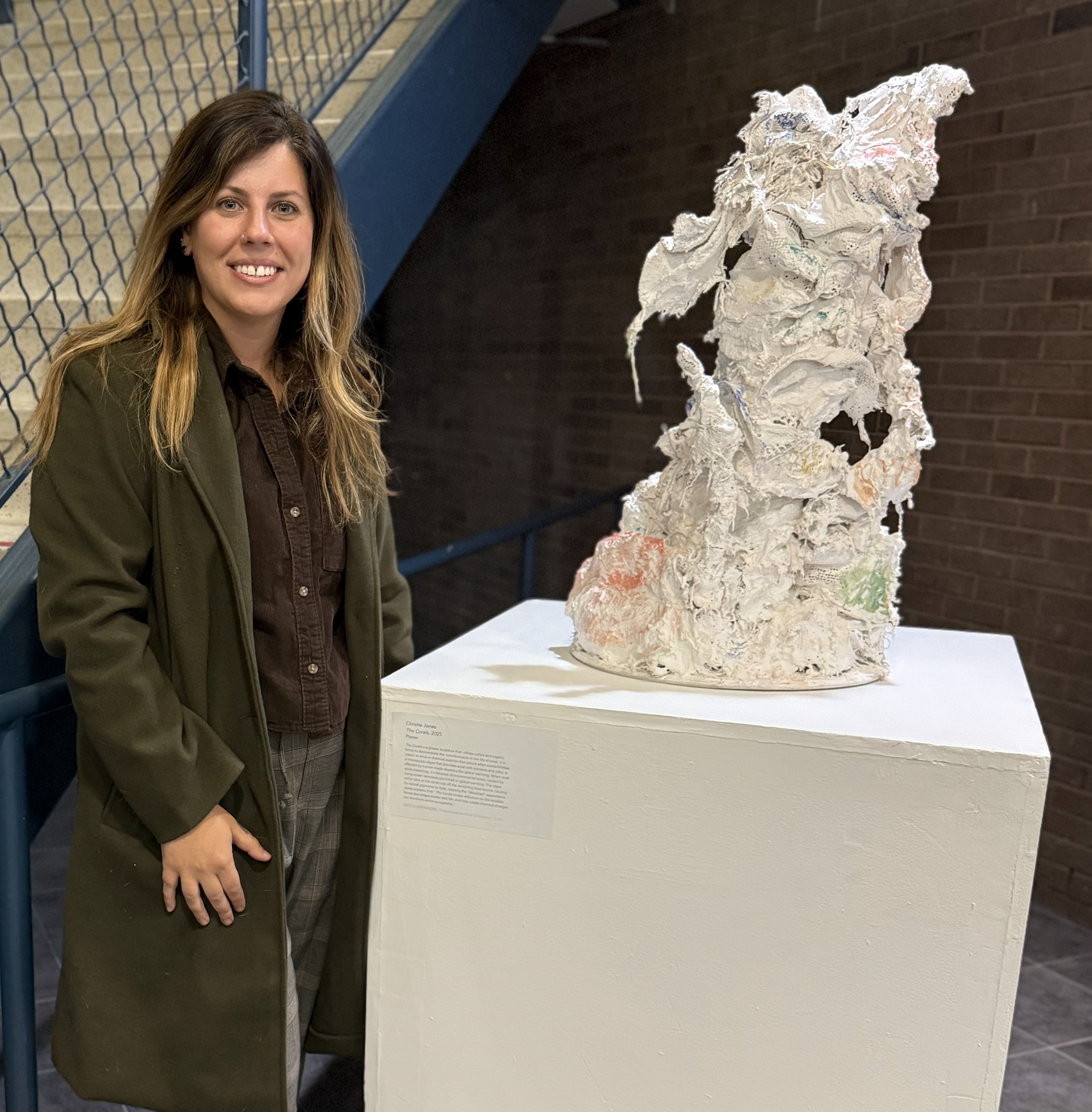 Individual wearing a dark green coat stands next to a plaster sculpture on a white pedestal  