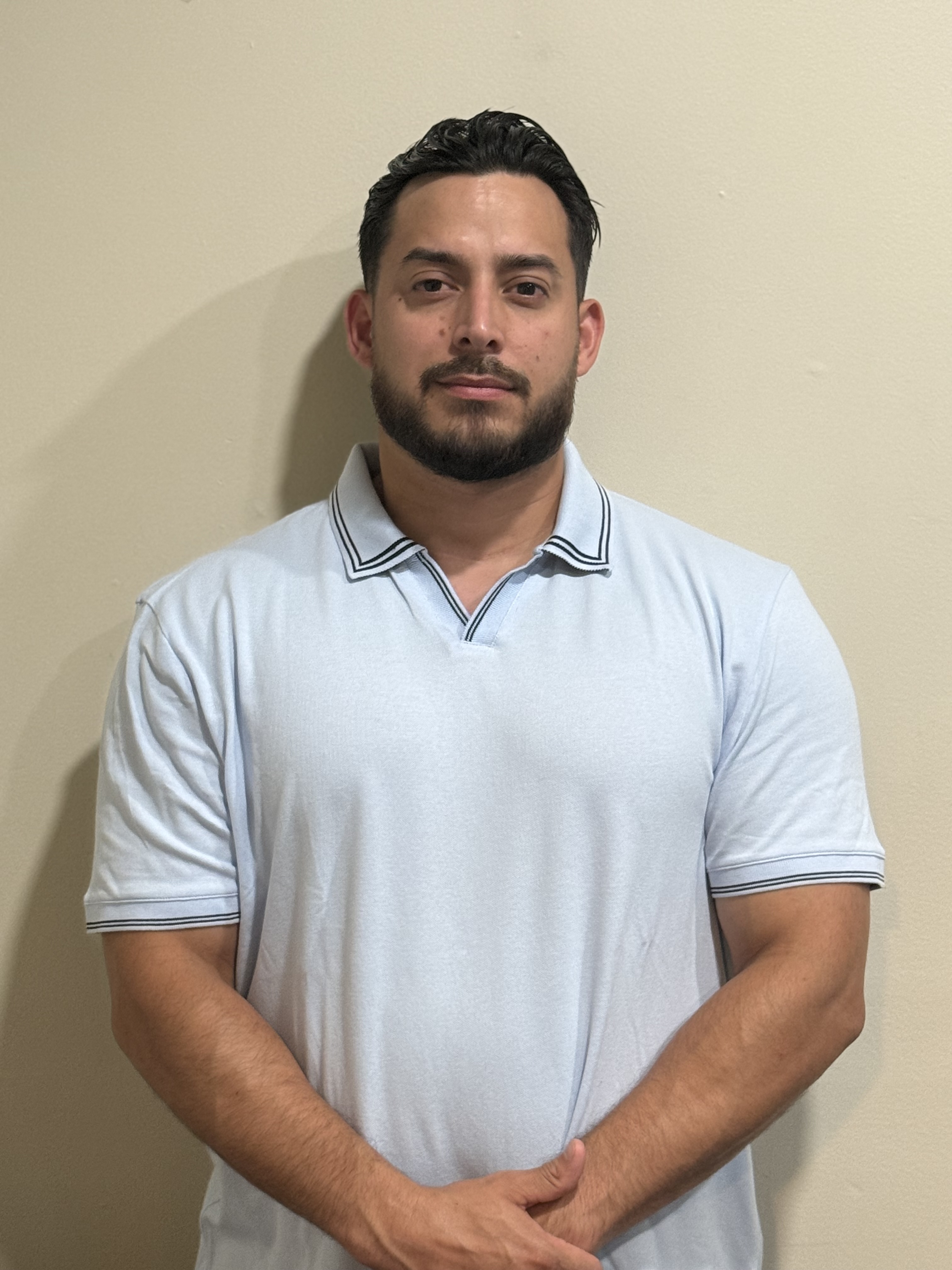 Individual in a light blue polo shirt standing against a neutral-colored wall.