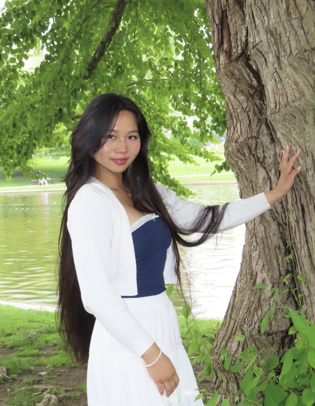 Person in a white outfit posing near a tree with a pond in the background.