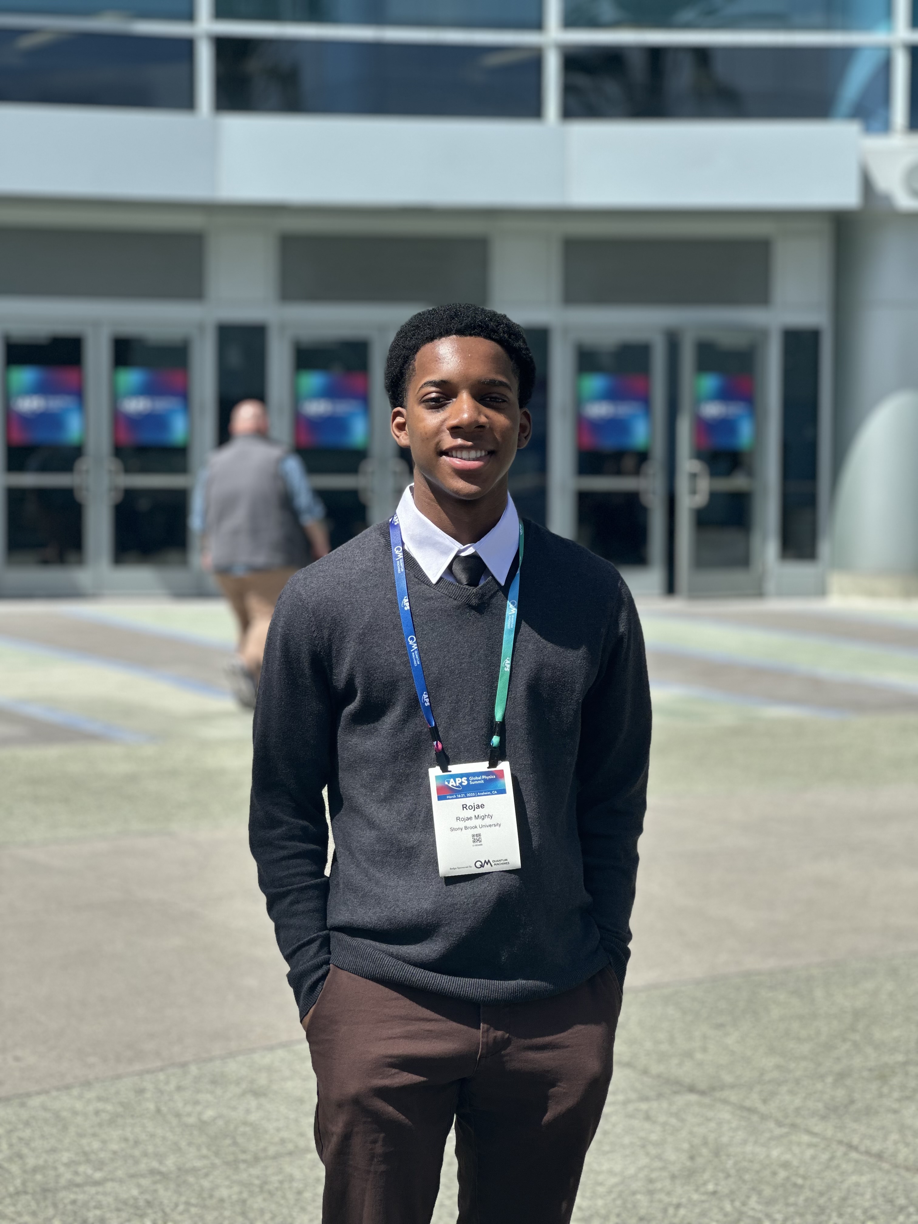 Person standing in front of a conference center wearing a badge and a smart outfit, smiling at the camera.