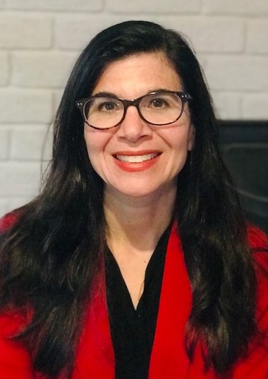 A woman with long dark hair and glasses smiles warmly, wearing a red jacket. She stands in front of a white brick wall, conveying a friendly and professional tone.