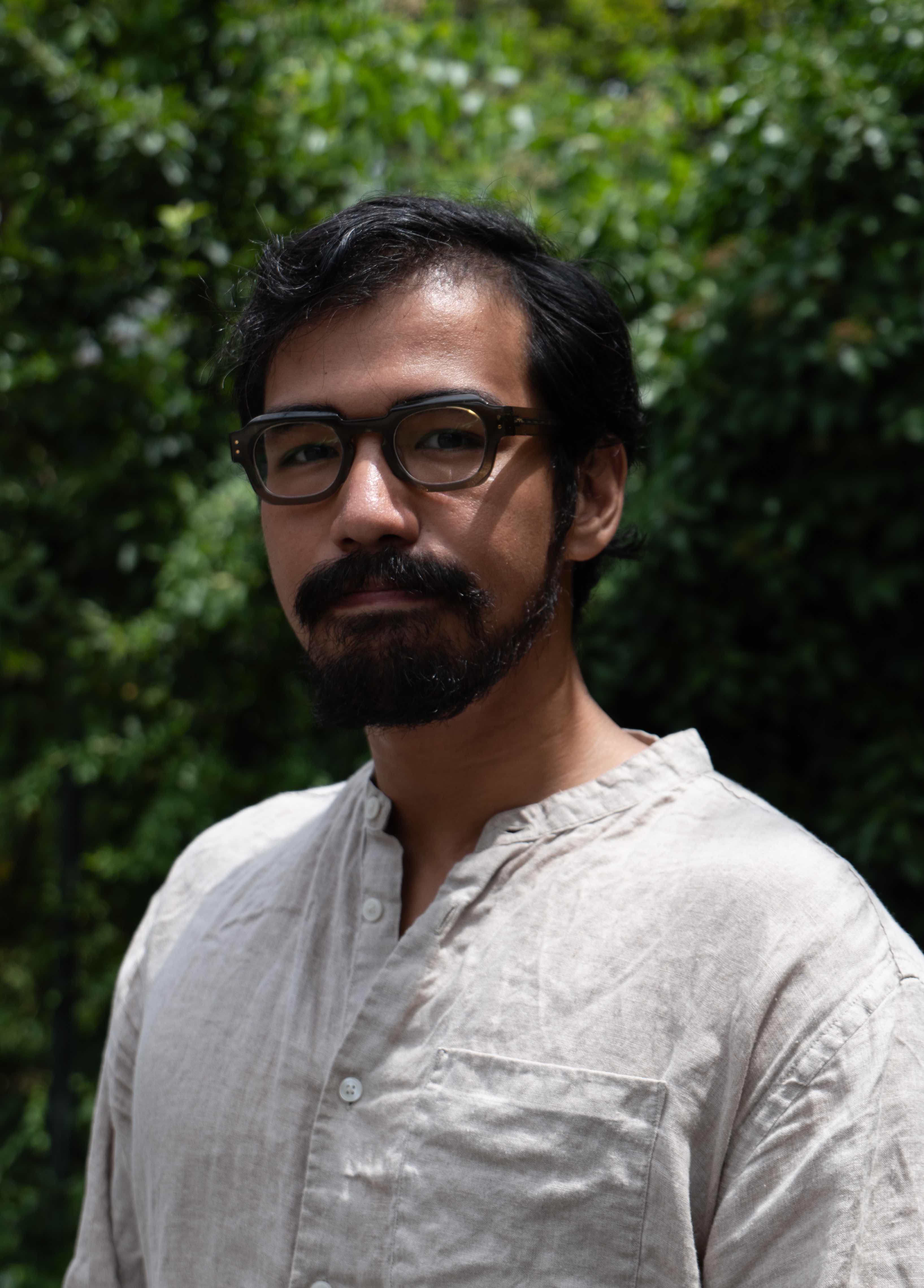 Person wearing glasses and a light beige shirt standing in front of a green, leafy background.