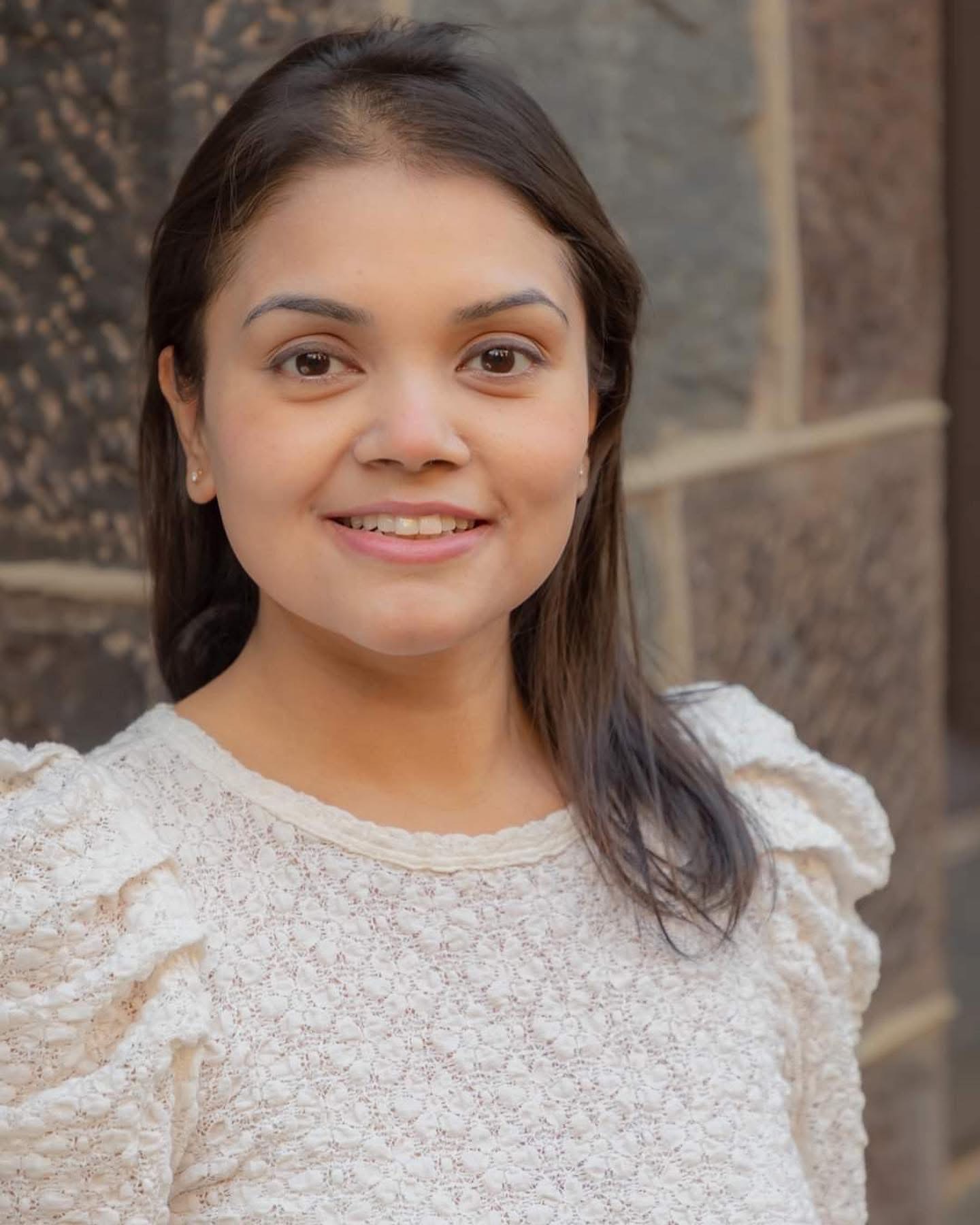 A person smiling at the camera, wearing a lace top, with a textured stone wall in the background.