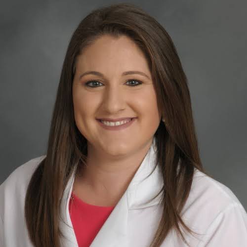 Portrait of a smiling healthcare professional in a white coat over a red top, set against a gray background.