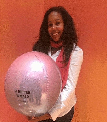 A person smiling and holding a large balloon with the phrase "A Better World" printed on it, against a vibrant orange background.