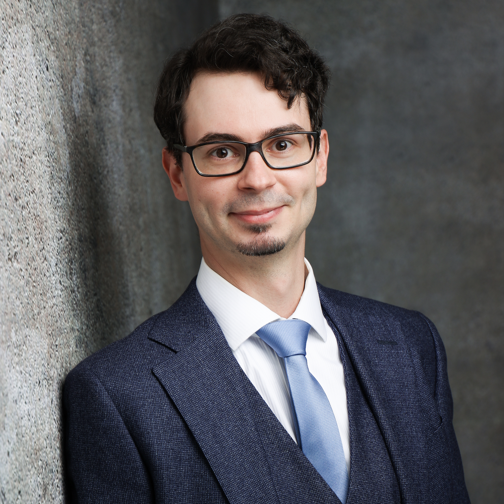 Portrait of a smiling individual in a formal blue suit and light blue tie, standing against a textured gray background.