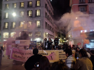 Colectivo Migrantes Members Protest in Valencia with people holding banners and signs, accompanied by smoke in the background.