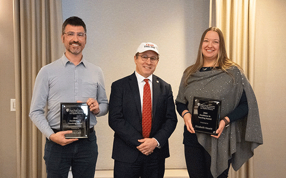Three people stand smiling in a room with neutral curtains. Two hold plaques, suggesting an award ceremony. The mood is celebratory and formal.