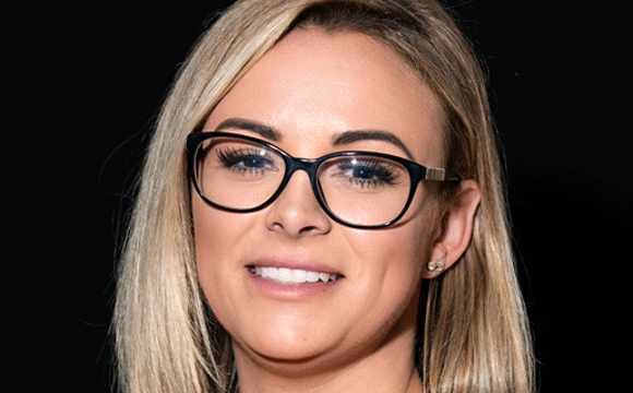 A woman with blonde hair and glasses smiles warmly against a dark background. Her expression is friendly, and she conveys a confident, approachable tone.
