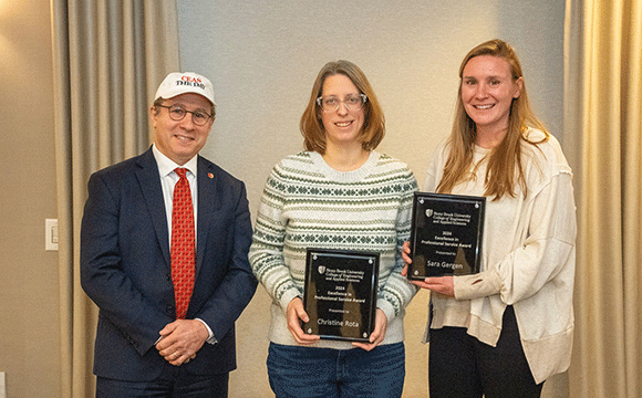 A man in a suit and red tie stands smiling next to two women holding plaques. The women are dressed casually, looking happy and proud in a warmly lit room.