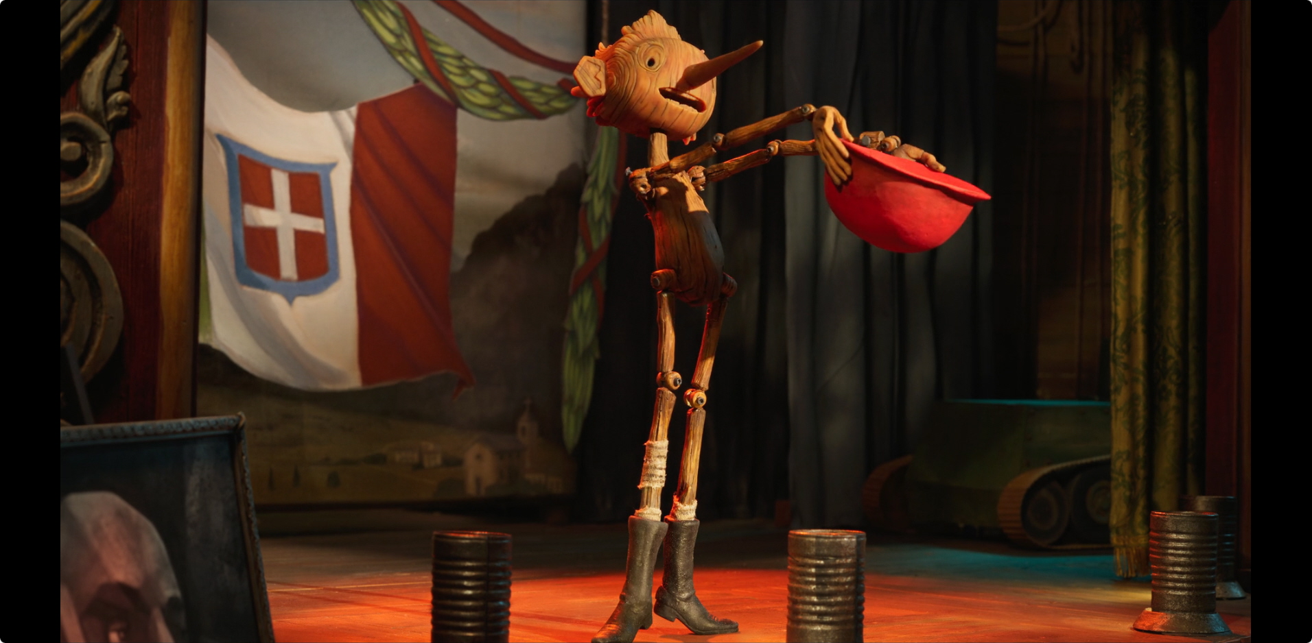 Guillermo del Toro's Pinocchio a screenshot from Guillermo del Toro's Pinocchio; Pinocchio is on a stage with the background being the flag of the kingdom of Italy
