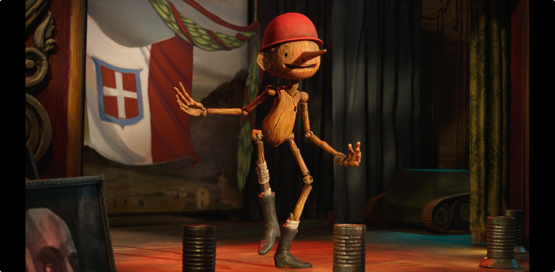 Guillermo del Toro's Pinocchio a screenshot from Guillermo del Toro's Pinocchio; Pinocchio is on a stage with the background being the flag of the kingdom of Italy