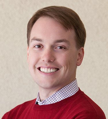 A head-and-shoulders portrait of a man with short brown hair and a bright, friendly smile. He is wearing a red crew-neck sweater over a light-colored patterned dress shirt. He is positioned against a neutral, off-white textured wall.