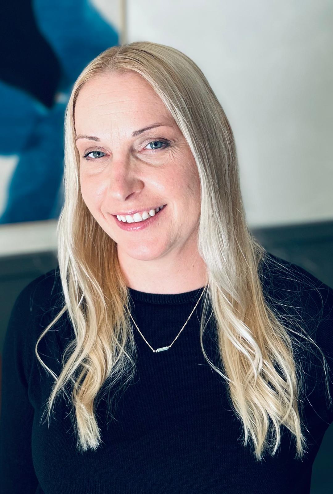Portrait-style image of a person with long blonde hair and a black top, wearing a pendant necklace. The background features abstract artwork in blue and white tones. 