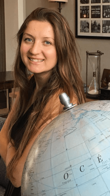 A portrait of a woman with long, wavy brown hair and blue eyes, smiling at the camera. She is leaning against a large globe of the Earth. The background is a warmly lit room featuring an hourglass and framed photos on the wall.
